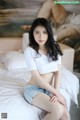 A woman sitting on a bed in a white shirt and denim shorts.